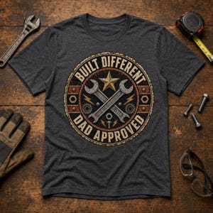 Peut inclure: Un t-shirt gris anthracite à manches courtes avec un motif circulaire portant le texte "BUILT DIFFERENT" et "DAD APPROVED". Le design comprend une étoile dorée centrale, deux clés croisées argentées, des engrenages, des boulons et des éclairs.