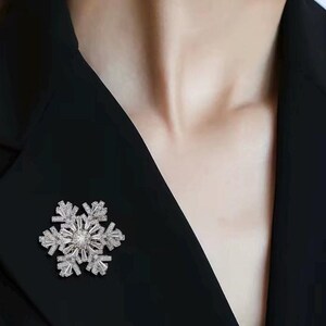 May include: A silver-toned snowflake brooch featuring a central round stone surrounded by multiple baguette and round cut clear crystals arranged in a symmetrical, six-pointed star pattern, pinned to a solid black fabric garment.