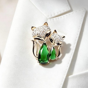 Puede incluir: Un broche dorado con forma de dos zorros, con detalles de cristales transparentes en las cabezas y grandes piedras de color verde esmeralda en forma de pera que forman los cuerpos. Las figuras de metal están pulidas sobre tela blanca.