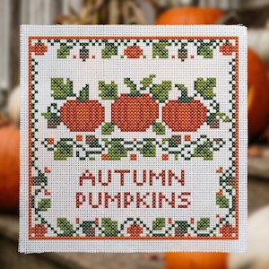 Può includere: Una tela quadrata per ricamo a punto croce con un motivo di tre zucche arancioni con foglie e viticci verdi. Il design include un bordo decorativo e il testo AUTUMN PUMPKINS in lettere arancioni su uno sfondo a griglia bianco.