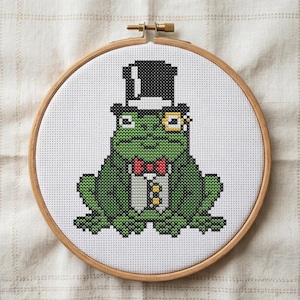 May include: Cross-stitch embroidery of a green frog wearing a black top hat, red bow tie, and monocle. The frog is dressed in a tuxedo with gold buttons. The embroidery is set in a wooden hoop.