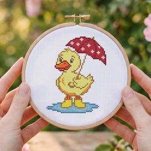 May include: Cross-stitch embroidery in a wooden hoop features a yellow duckling wearing yellow boots and holding a red umbrella with white crosses. The duckling stands in a blue puddle. The background is blurred with green and pink foliage.