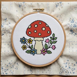 May include: A cross-stitch embroidery hoop featuring a red mushroom with white spots, surrounded by colorful flowers and green leaves. The design is set against a white background and framed by a wooden hoop. The embroidery is on a cream-colored fabric.