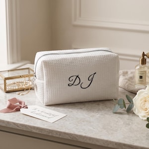 Könnte beinhalten: Weiße Kosmetiktasche mit Waffelstruktur und schwarz gestickten Initialen "DJ". Die Tasche hat einen schwarzen Reißverschluss und ein Etikett mit der Aufschrift "To a beautiful bridesmaid". Eine goldgerahmte Schmuckschatulle und eine Parfümflasche sind ebenfalls zu sehen.