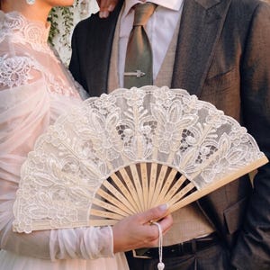 May include: A cream lace fan with a floral pattern is held. The fan has a wooden frame and a white tassel. The image also shows a person in a white dress and a person in a suit.