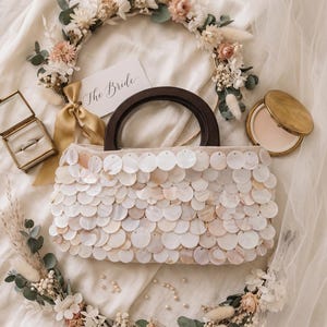 May include: A cream-colored handbag adorned with circular shell accents, featuring a dark brown wooden handle. A floral wreath and a compact mirror are nearby. A card reads "The Bride". A ring is in a small glass box.