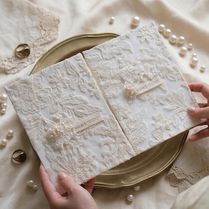 Puede incluir: Dos libros blancos de votos matrimoniales con intrincados diseños de encaje floral. Cada libro tiene una pequeña etiqueta beige que dice "VOWS". Los libros están sobre una bandeja dorada, rodeados de perlas y encaje.