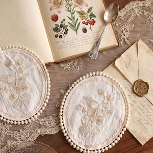 Puede incluir: Dos posavasos bordados blancos con diseños florales y un ribete de pompón. Un libro abierto con ilustraciones botánicas, una cuchara de plata y una carta vintage con un sello de cera también son visibles.