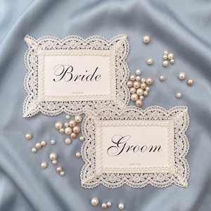May include: Two white lace wedding place cards with the words "Bride" and "Groom" in elegant script. The cards are surrounded by pearls and set on a light blue satin background. The date "Jan 10 2036" is printed on the cards.