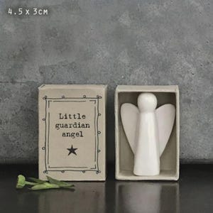 Puede incluir: Una pequeña figura de ángel de cerámica blanca dentro de una caja tipo cerilla. La caja es de color beige con las palabras "Little guardian angel" y una estrella. El ángel mide aproximadamente 4,5 x 3 cm.