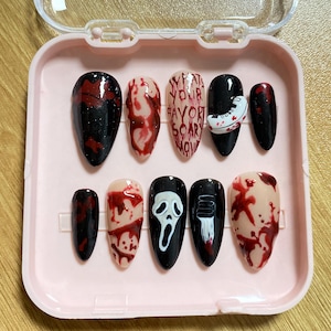 May include: A set of ten press-on nails in a pink case. The nails feature various designs, including blood splatter, a ghost face, and the text "WHAT'S YOUR FAVORITE SCARY MOVIE?" in a spooky font. The nails are black, red, and nude.