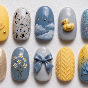 May include: A collection of ten artificial nails with various designs. The designs include a yellow duck feet, speckled patterns, clouds with flying birds, a yellow duck, glitter, floral patterns, a blue bow, a knitted pattern, and a blue flower.