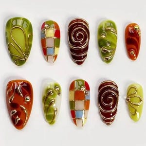 May include: A collection of ten artificial nails in various designs. The nails feature a range of colors, including green, orange, red, and yellow, with gold wire and jewel accents. Some nails have a swirl design, while others have a checkered pattern.
