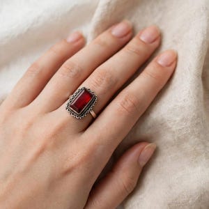 May include: A silver ring featuring a rectangular, deep red gemstone set in a decorative, scalloped metal frame. The band is thin and metallic, showcasing a polished finish that complements the dark, vibrant color of the central stone setting.