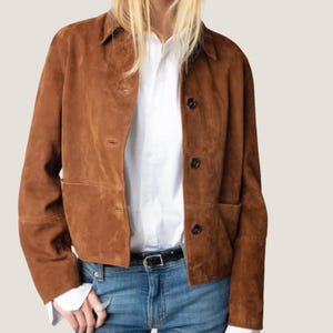 May include: A brown suede jacket with a button-up front, two front pockets, and a collared neckline. The jacket is worn over a white button-down shirt and blue jeans. The model is also wearing a black belt.
