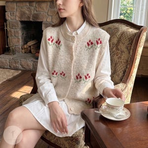 Puede incluir: Un chaleco de punto color crema con botones y bordado floral rojo. El chaleco se lleva sobre una camisa blanca con cuello. La imagen muestra a una persona sentada en un sillón estampado, sosteniendo una taza y un platillo.