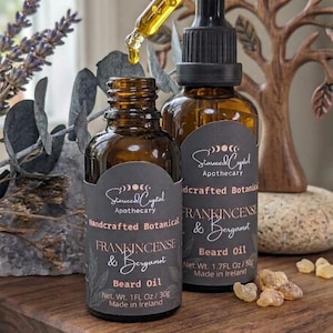 Op de afbeelding: Twee amberkleurige glazen flessen Starseed Crystal Apothecary Frankincense & Bergamot baardolie met zwarte druppeldop. Eén fles is geopend met een glazen pipet die goudkleurige olie afgeeft. Etiketten vermelden 30g en 50g, gemaakt in Ierland.