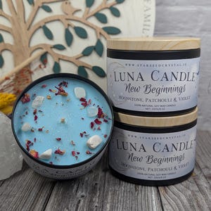 May include: A light blue candle with decorative crystals and dried flower petals sits next to two stacked Luna Candles. The candles are in black tins with wooden lids. The text on the label reads "Luna Candle New Beginnings".