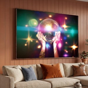 Puede incluir: Una impresión de arte mural rectangular que muestra una bola de discoteca sostenida por dos manos sobre un fondo oscuro con destellos de luz vibrantes y multicolores y efectos bokeh en tonos verde, rosa, amarillo y azul, creando una atmósfera brillante.