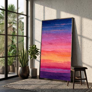 Puede incluir: Una impresión artística enmarcada que representa una escena abstracta de atardecer. La obra de arte muestra un degradado de colores, incluyendo tonos morados profundos, naranja, rosa y azul. La impresión se exhibe en una habitación con luz natural, plantas y un taburete de madera.