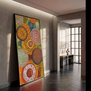 Puede incluir: Gran impresión artística abstracta enmarcada con círculos concéntricos coloridos en tonos naranja, amarillo, verde y marrón. La obra de arte se apoya contra una pared gris en un entorno interior moderno. La impresión tiene un marco negro.