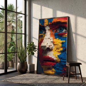 Puede incluir: Una obra de arte enmarcada que representa un rostro humano construido con ladrillos de juguete de colores. El retrato presenta ojos azules, una nariz blanca y labios rojos, con un fondo de ladrillos amarillos, azules y rojos. La obra se exhibe en una habitación con luz natural.