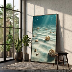 May include: A framed art print showing an underwater scene with sunlight filtering through the water. The image shows a sandy seabed with scattered seashells. The artwork is displayed in a room with plants and a window.