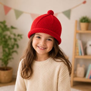 Può includere: Un berretto in maglia rosso brillante con bordo arrotolato e un piccolo pompon sulla parte superiore. Il cappello presenta una consistenza a coste ed è indossato sopra capelli lunghi castani. È realizzato in lana con una forma arrotondata.