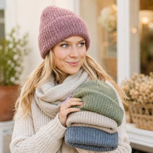 May include: A dusty rose knit beanie worn on the head, paired with a beige scarf and a cream sweater. Three additional folded knit beanies in olive green, beige, and slate blue are held in the hands, showcasing a ribbed texture and cuff.