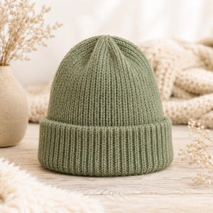 Puede incluir: Un gorro de punto color verde salvia con una textura de canalé vertical gruesa. El gorro presenta un borde ancho doblado en la base y una corona redondeada, confeccionado con un patrón de punto consistente en toda la prenda para un aspecto uniforme.