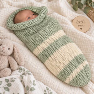 Puede incluir: Un bebé envuelto en un capullo de punto a rayas de color verde salvia y crema. Se ven los ojos y la frente del bebé. Un disco de madera redondo tiene grabado el texto little one y un corazón. Un conejo de peluche está colocado cerca.