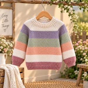 Puede incluir: Un suéter de punto de manga larga con rayas horizontales en crema, lavanda, verde salvia, melocotón y rosa empolvado. La prenda tiene un cuello redondo acanalado, puños acanalados y un dobladillo acanalado, colgado en una percha de madera.