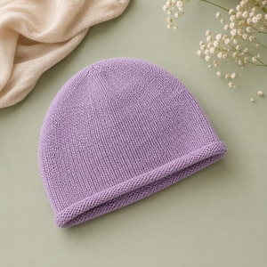 May include: A solid light purple knit beanie with a rounded crown and a thick, rolled-up brim. The hat features a consistent stockinette stitch pattern throughout the main body, providing a textured appearance and a simple, clean design structure.
