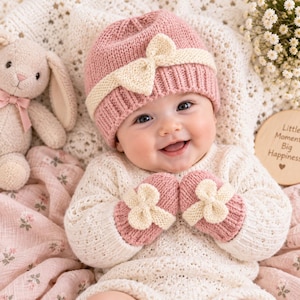 Könnte beinhalten: Eine altrosa Strickmütze mit cremefarbener Schleife und dazu passende Strickfäustlinge, die jeweils mit einer cremefarbenen Schleife verziert sind. Das Set wird mit einem cremefarbenen Langarmpullover mit durchgehendem, strukturiertem Strickmuster getragen.