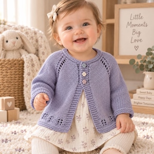 May include: A light purple knitted cardigan featuring long sleeves, a round neckline, and a button-front closure with three small flower-shaped buttons. The garment displays a delicate eyelet pattern along the hem and raglan sleeve seams.