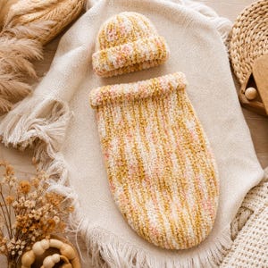 Puede incluir: Un saco de dormir tipo capullo para bebé, suave y afelpado, junto con un gorro a juego, ambos confeccionados con hilo jaspeado en tonos amarillo mostaza, crema y rosa suave. El conjunto presenta un patrón de punto acanalado y texturizado.