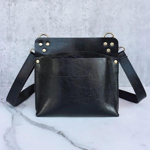 May include: Black leather barber tool bag with a shoulder strap. The bag has a square shape with a large front pocket and brass-colored rivets. The top of the bag has two brass-colored rings for the shoulder strap to attach to.