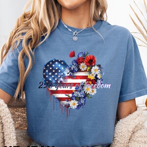 May include: Blue jean colored t-shirt with a heart-shaped design featuring the American flag and flowers. The text "250 years of freedom" is printed on the heart. The shirt is a comfortable, casual style.