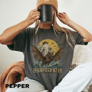 May include: A charcoal gray t-shirt featuring a graphic of a ghost in a cowboy hat riding a large bat over ocean waves in front of a yellow moon, with bats flying nearby and the text "HIGHFIELD 87 CO." printed in a bold, serif font below.