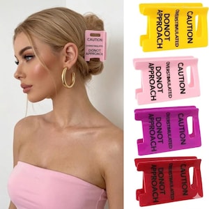 Könnte beinhalten: Ein Set aus vier rechteckigen Haarspangen in Gelb, Hellrosa, Lila und Rot. Jede Spange trägt den schwarzen Aufdruck CAUTION OVERSTIMULATED DO NOT APPROACH. Die Spangen haben ein ausgeschnittenes Design und werden einzeln sowie im Gebrauch gezeigt.