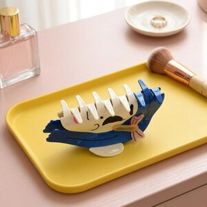 May include: A whimsical hair claw clip in the shape of a cartoon character, featuring white, blue, and pink accents. The clip is set on a yellow tray, with a perfume bottle and makeup brush in the background. This accessory is perfect for adding a playful touch to any hairstyle.