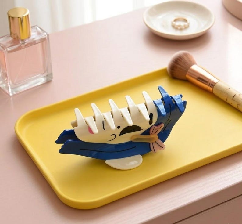 May include: A whimsical hair claw clip in the shape of a cartoon character, featuring white, blue, and pink accents. The clip is set on a yellow tray, with a perfume bottle and makeup brush in the background. This accessory is perfect for adding a playful touch to any hairstyle.