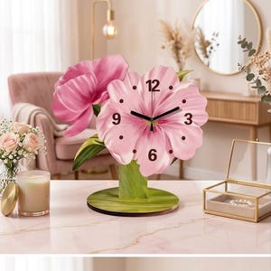 May include: A tabletop clock shaped like a large pink flower with green leaves and a stem base. The clock face features black numbers at the 12, 3, 6, and 9 positions, with black hour, minute, and second hands centered on the pink petals.
