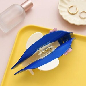 May include: A blue and white hair clip in the shape of a dolphin, with a gold-colored spring mechanism. The clip is on a yellow tray, with a perfume bottle and a small dish with gold earrings in the background.