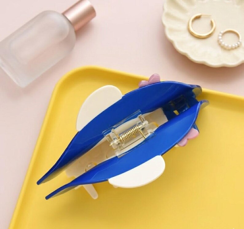 May include: A blue and white hair clip in the shape of a dolphin, with a gold-colored spring mechanism. The clip is on a yellow tray, with a perfume bottle and a small dish with gold earrings in the background.