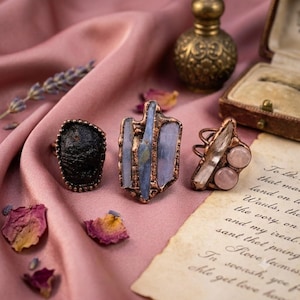 May include: Three rings with copper-toned metal settings. The left ring features a dark, textured stone. The center ring displays three vertical, light blue crystal shards. The right ring holds a clear crystal point and two round, pale pink stones.