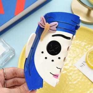 May include: A blue and white decorative hair clip in the shape of a stylized face, featuring blue hair, a black button eye, and pink lips. A pink and yellow butterfly adorns the top. The clip is held by a hand.