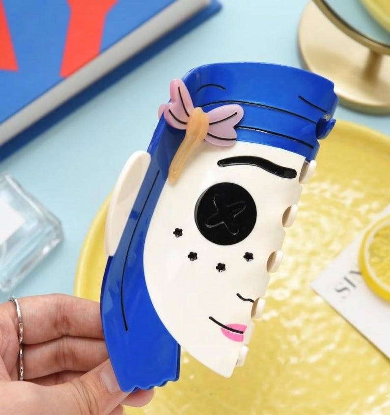 May include: A blue and white decorative hair clip in the shape of a stylized face, featuring blue hair, a black button eye, and pink lips. A pink and yellow butterfly adorns the top. The clip is held by a hand.