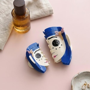May include: Two blue and white face-shaped hair clips with pink accents. One perfume bottle with a wooden cap is visible. A small ceramic dish with gold earrings is also present. The items are on a pink surface.