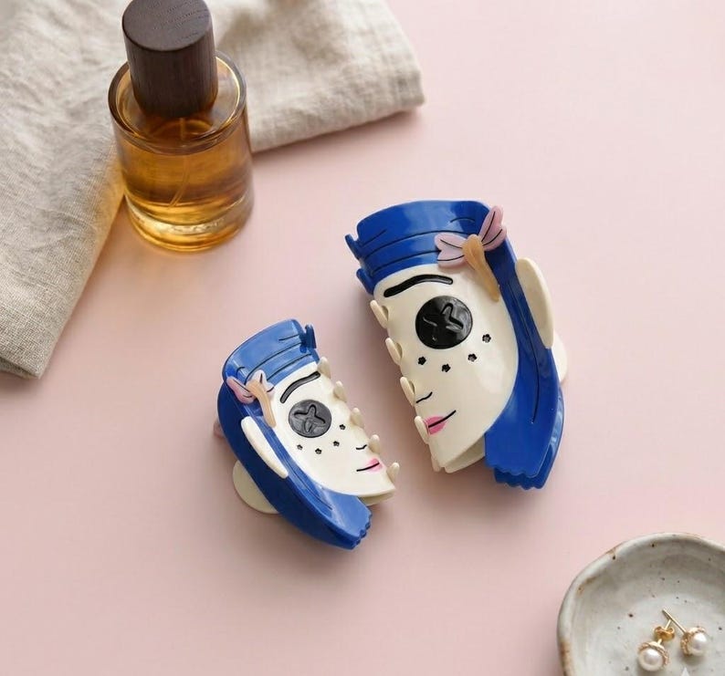 May include: Two blue and white face-shaped hair clips with pink accents. One perfume bottle with a wooden cap is visible. A small ceramic dish with gold earrings is also present. The items are on a pink surface.